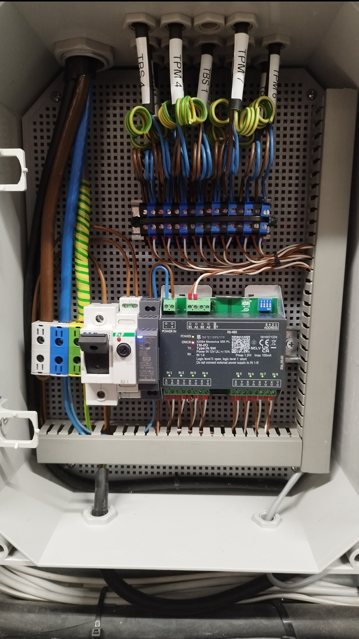 Otwarta skrzynka elektryczna z widocznymi przewodami, zabezpieczeniami, listwami zaciskowymi i modułem sterującym z oznaczeniami, starannie ułożone kable wewnątrz szarej obudowy.