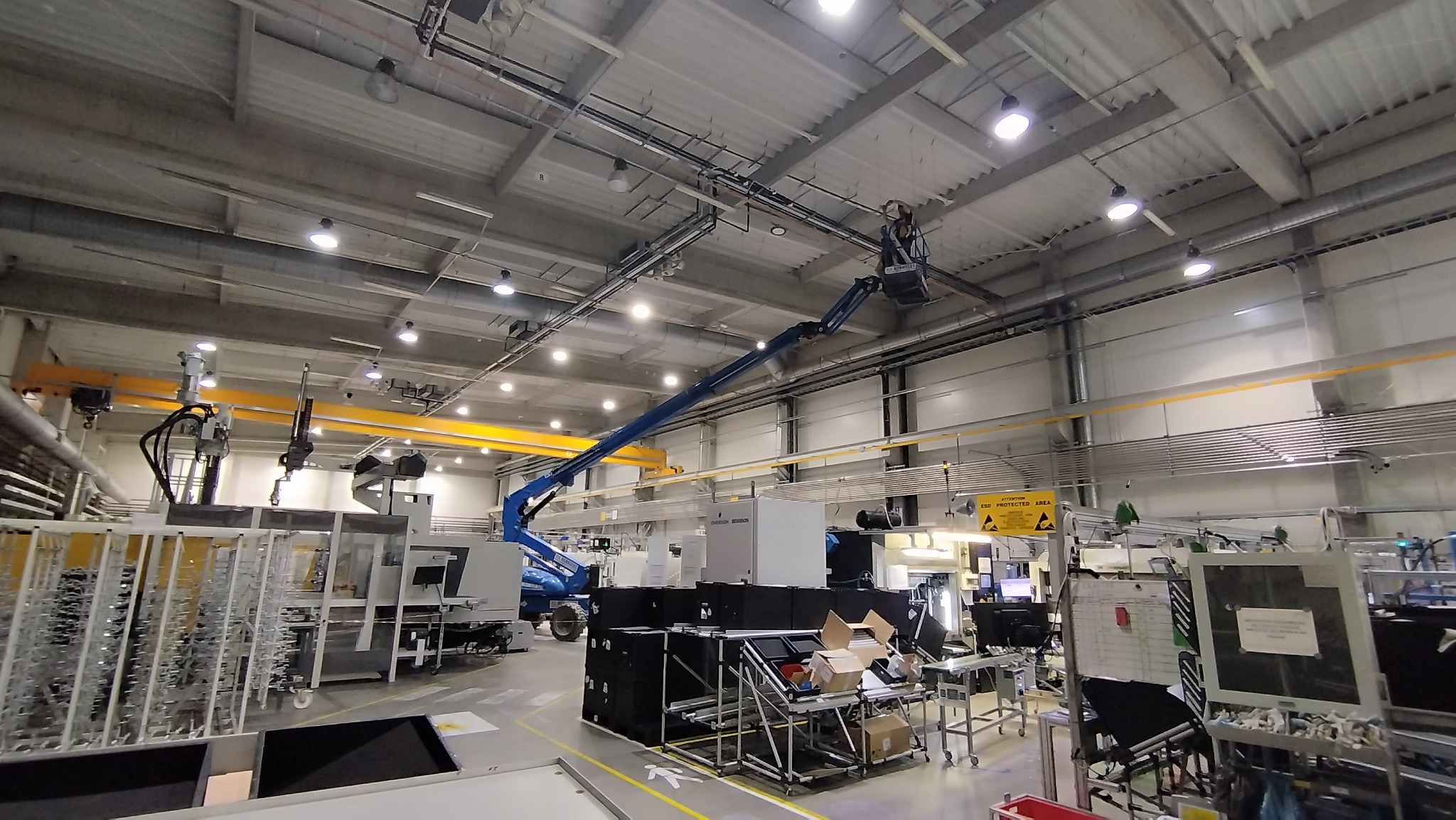 Pracownik na podnośniku koszowym wykonuje prace na wysokości pod sufitem hali przemysłowej, widoczne rury i przewody pod stropem, w tle maszyny i urządzenia produkcyjne.