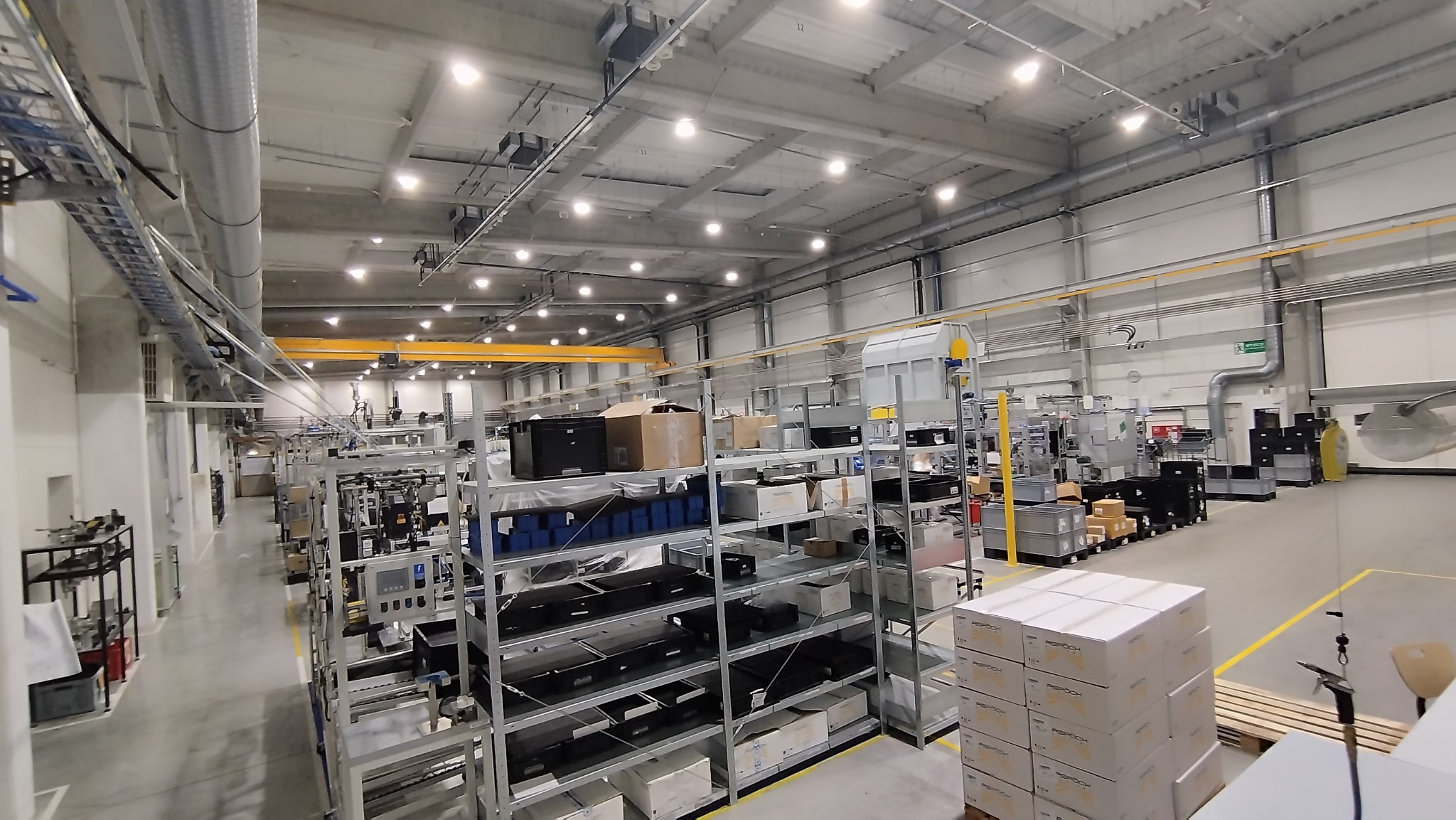 Przestronna hala produkcyjna z regałami wypełnionymi komponentami, widoczne oświetlenie sufitowe i instalacje wentylacyjne, suwnica pod stropem