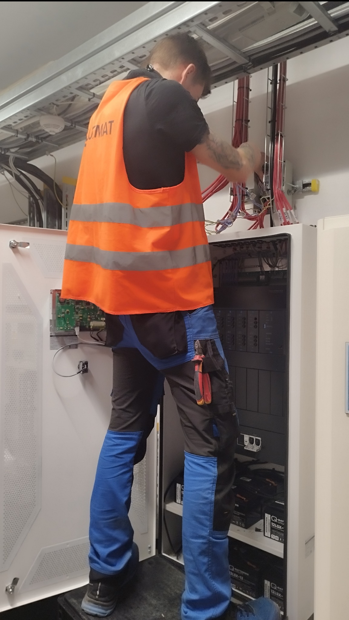 Elektryk w pomarańczowej kamizelce roboczej, stojący na podwyższeniu, pracuje nad podłączeniem przewodów wewnątrz białej szafy elektrycznej z widocznymi akumulatorami i modułami sterującymi,...