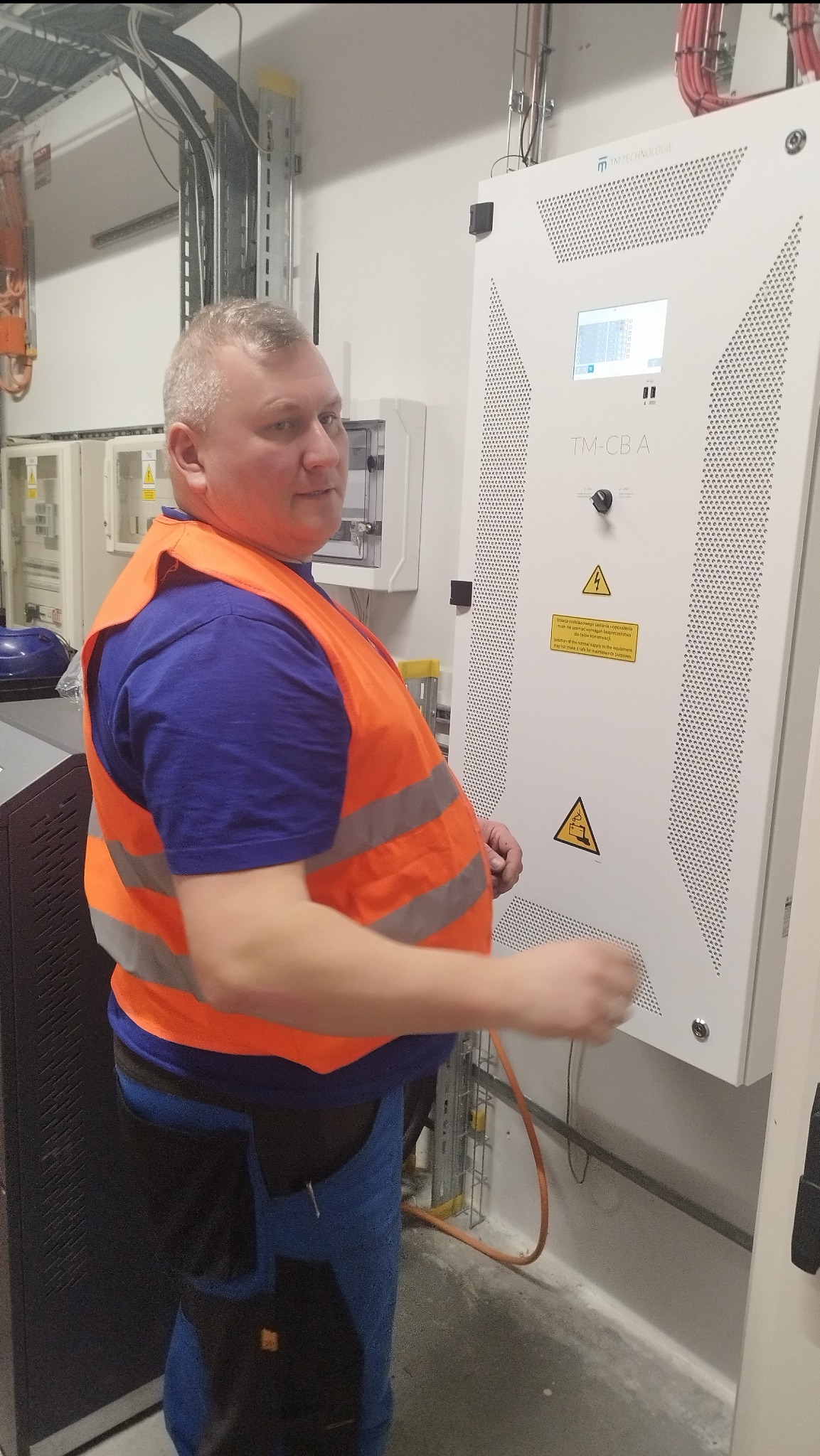 Elektryk w pomarańczowej kamizelce odblaskowej stoi obok białej szafy sterowniczej z wyświetlaczem i ostrzeżeniami o napięciu, w tle widoczne kable i inne urządzenia elektryczne.