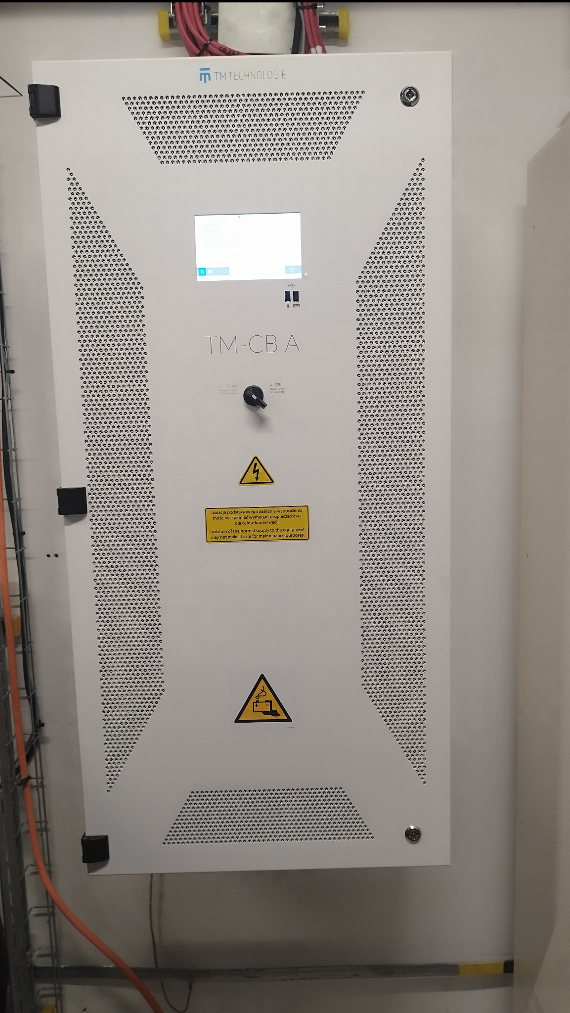 Biała szafa sterownicza TM Technologie TM-CB A z perforowanymi panelami, wyświetlaczem, przełącznikiem i żółtymi ostrzeżeniami o napięciu, zamontowana na ścianie w pomieszczeniu technicznym.