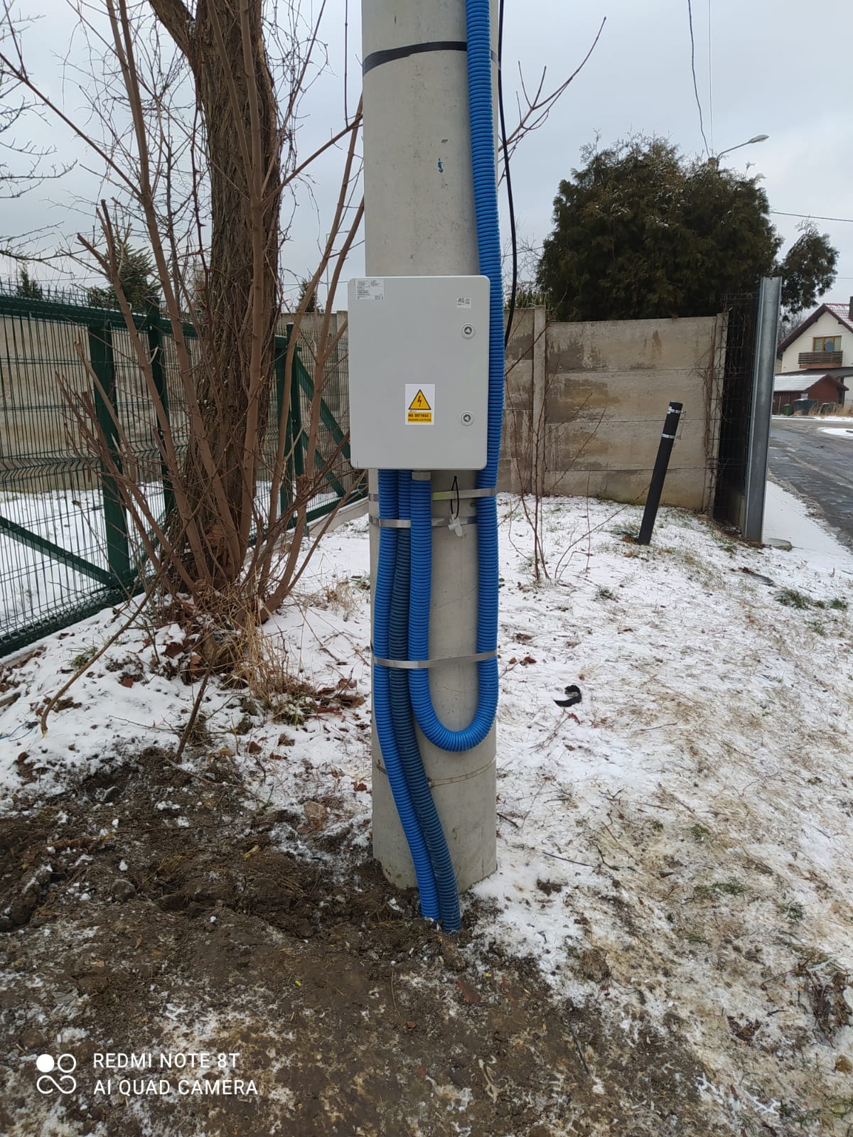 Szara skrzynka elektryczna z symbolem ostrzegawczym na betonowym słupie, zabezpieczona niebieskimi rurami karbowanymi, na zewnątrz w zimowej scenerii.