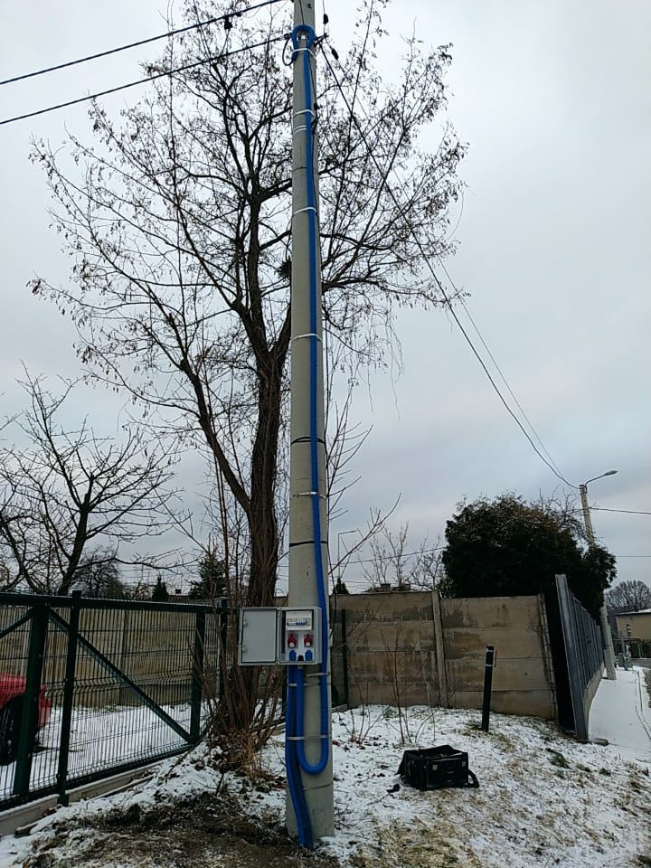 Słup energetyczny z zamontowaną skrzynką elektryczną i niebieskimi rurami ochronnymi w zimowym krajobrazie, widoczne ogrodzenie i drzewo bez liści.