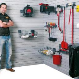 Debar system - Mężczyzna oparty o ścianę z systemem przechowywania narzędzi i akcesoriów w garażu lub pomieszczeniu gospodarczym, widoczne kosze, wieszaki i różne narzędzia.
