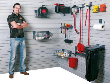 Mężczyzna oparty o ścianę z systemem przechowywania narzędzi i akcesoriów w garażu lub pomieszczeniu gospodarczym, widoczne kosze, wieszaki i różne narzędzia.