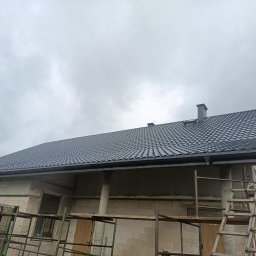 Pro-dom budownictwo - Szary dach z blachy trapezowej na domu w stanie surowym, widoczne rusztowania i okna do wstawienia, pochmurne niebo w tle.