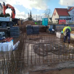 Zbrojenie fundamentów pod budynek jednorodzinny, widoczny pracownik w odblaskowej kamizelce, teren budowy z dźwigiem i przenośną toaletą w tle.
