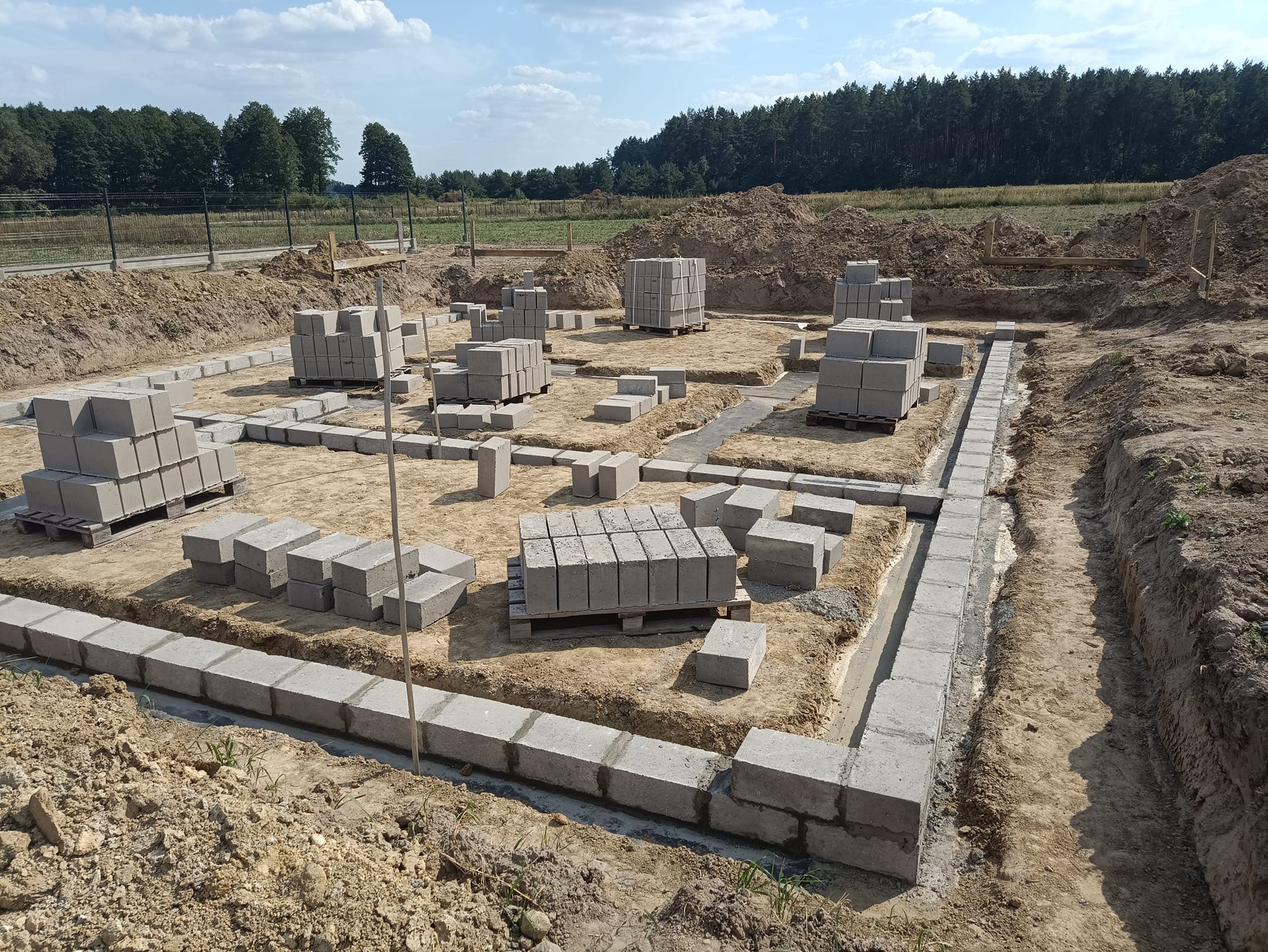 Wykopane fundamenty z częściowo ułożonymi bloczkami betonowymi na zaprawie, palety z bloczkami w tle, widok na pole i las.