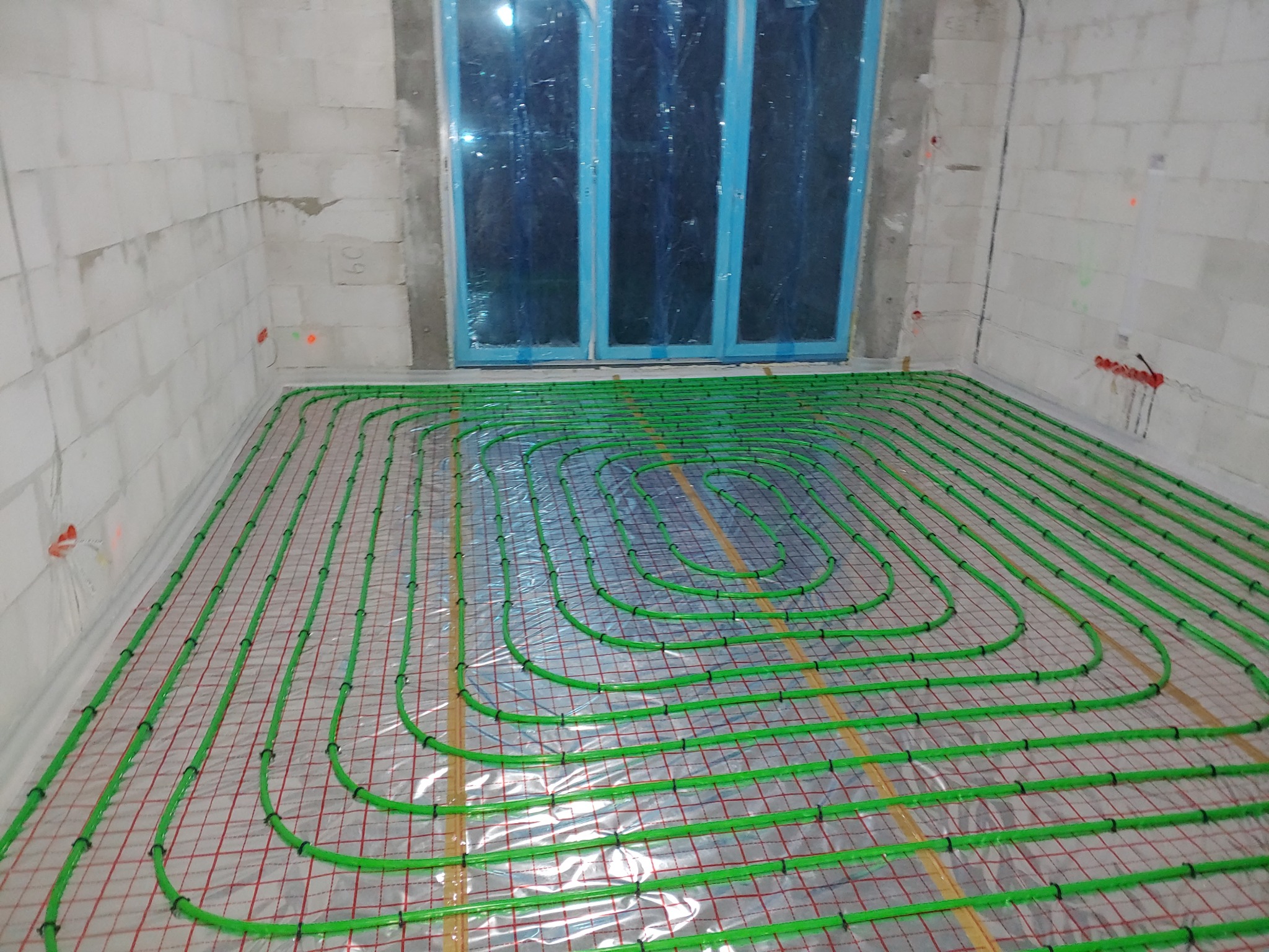 Instalacja ogrzewania podłogowego z zielonych rurek na folii z czerwoną kratką w surowym pomieszczeniu z oknem zabezpieczonym folią.