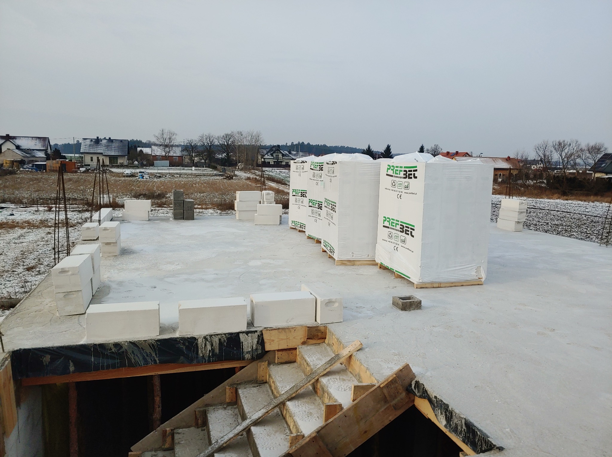 Fundamenty domu w trakcie budowy, widoczne bloczki betonowe na płycie fundamentowej, palety z materiałem budowlanym Prefbet w folii oraz drewniane schody do piwnicy, w tle zabudowa jednorodzinna...