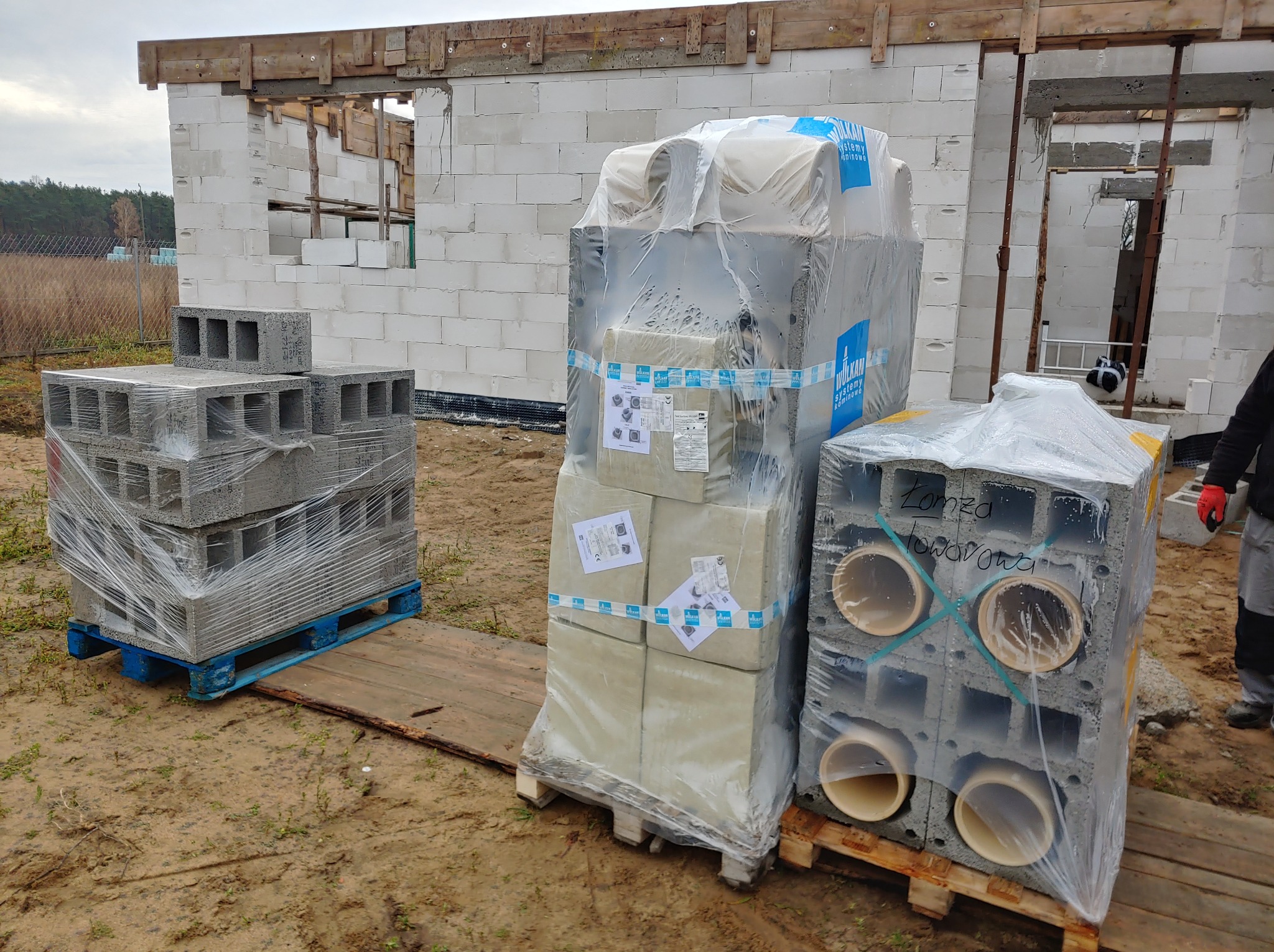 Świeżo dostarczone, zafoliowane bloczki betonowe i pustaki wentylacyjne na paletach, składowane przy budowie domu z białego betonu komórkowego.