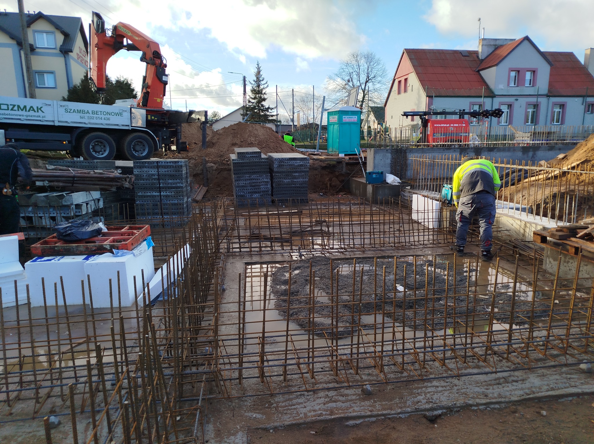 Zbrojenie fundamentów pod budynek jednorodzinny, widoczny pracownik w odblaskowej kamizelce, teren budowy z dźwigiem i przenośną toaletą w tle.