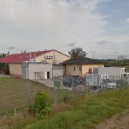 Widok z zewnątrz na budynki gospodarcze z rampą załadunkową, zaparkowanymi samochodami dostawczymi i ciężarówkami, ogrodzone siatką.