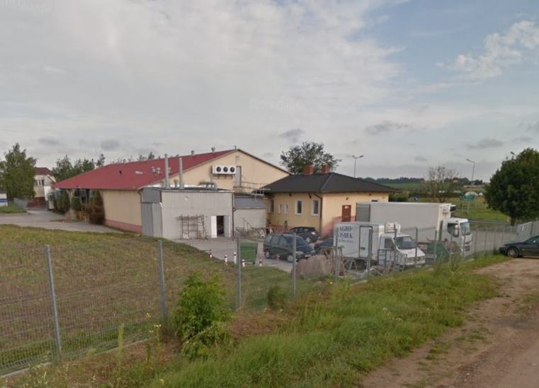 Widok z zewnątrz na budynki gospodarcze z rampą załadunkową, zaparkowanymi samochodami dostawczymi i ciężarówkami, ogrodzone siatką.