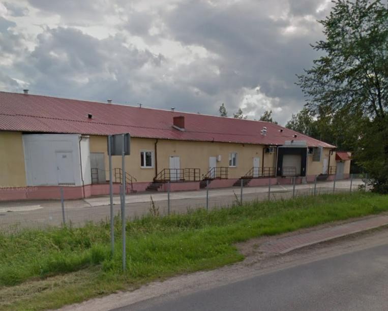 Budynek magazynowy z rampami załadunkowymi, czerwonym dachem i jasną elewacją, otoczony niskim ogrodzeniem i zielenią.