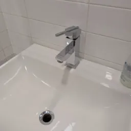 Błyszcząca, chromowana bateria umywalkowa na czystej, białej umywalce w łazience po sprzątaniu w Warszawie. Widoczne białe płytki i dozownik mydła.