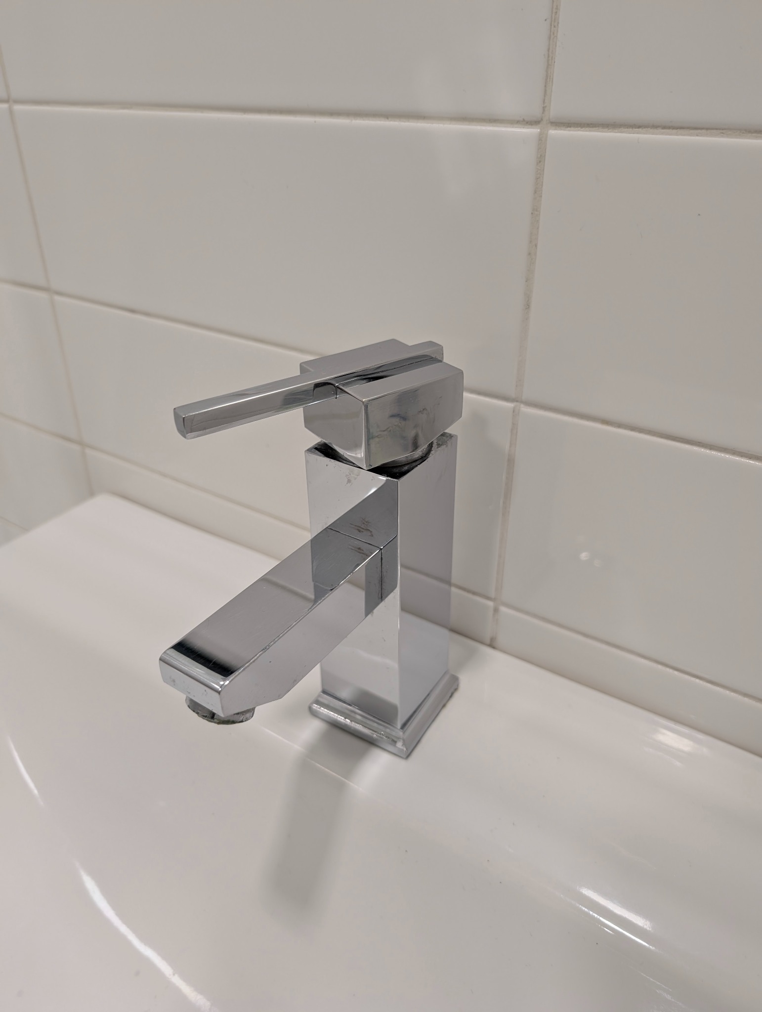 Błyszcząca, chromowana bateria umywalkowa w łazience z białymi płytkami. Widoczne ślady użytkowania na baterii i fugach.