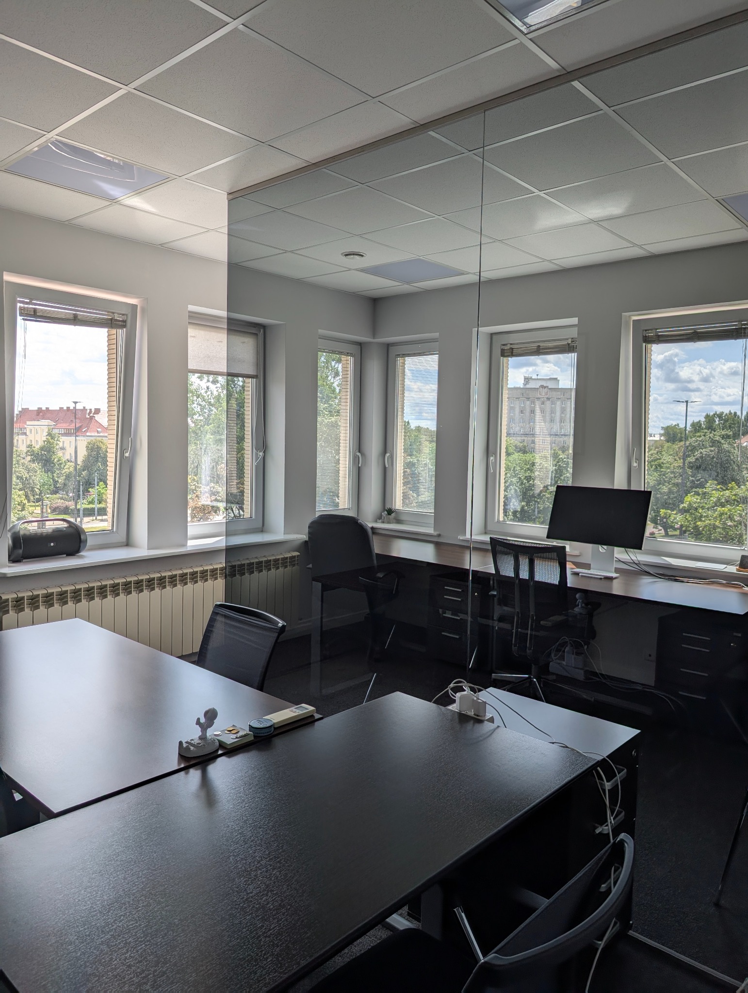 Nowoczesne biuro z przeszkloną ścianą działową, biurkami, krzesłami i widokiem na park przez okna z żaluzjami. Na biurkach akcesoria biurowe i kable.