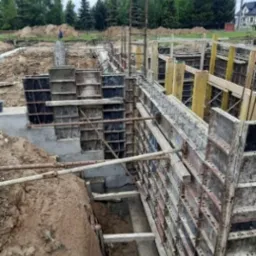 Budowa fundamentów: szalunki ustawione w wykopie, widoczne zbrojenie, przygotowanie do wylewania betonu. Tło z drzewami i domami.