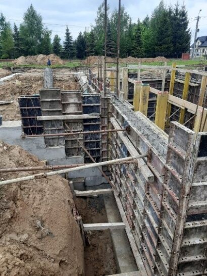 Budowa fundamentów: szalunki ustawione w wykopie, widoczne zbrojenie, przygotowanie do wylewania betonu. Tło z drzewami i domami.