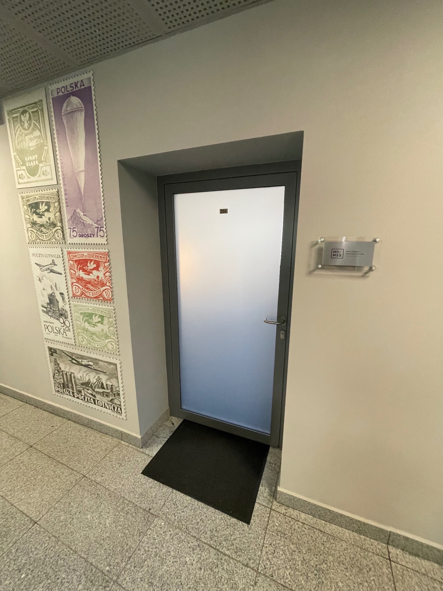 Wejście do biura z matową szybą i szyldem MD MSS, obok ściana zdobiona znaczkami pocztowymi, czarna wycieraczka przed drzwiami, szare płytki na podłodze.