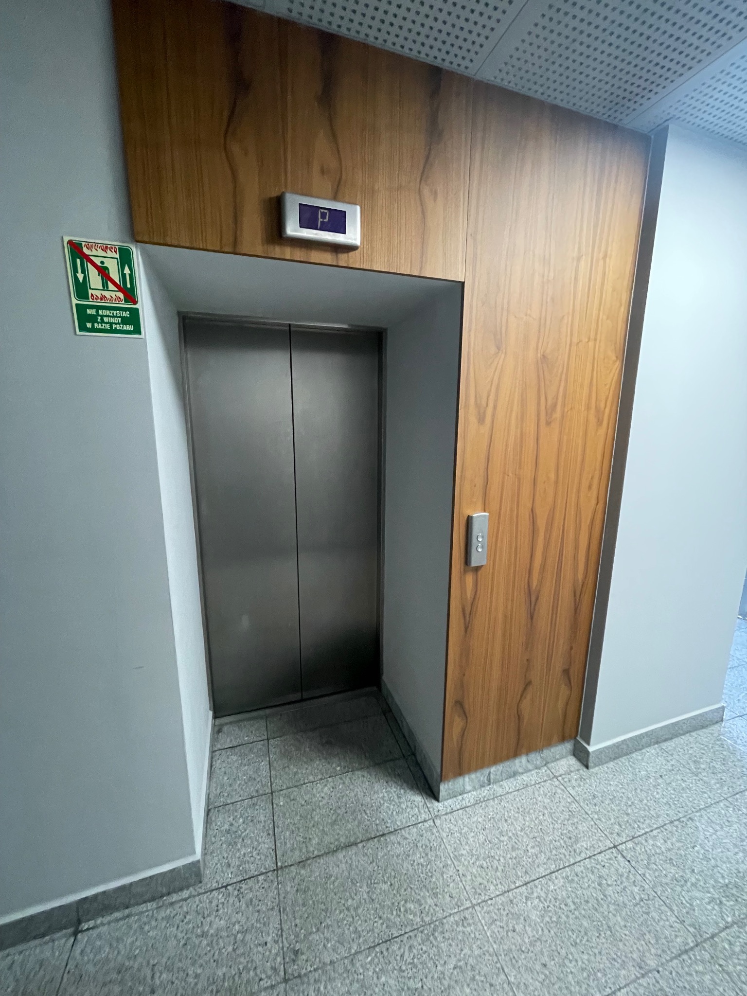 Metalowe drzwi windy w drewnianej obudowie w biurowcu. Na ścianie znak zakazu używania windy w razie pożaru. Przycisk windy po prawej stronie.