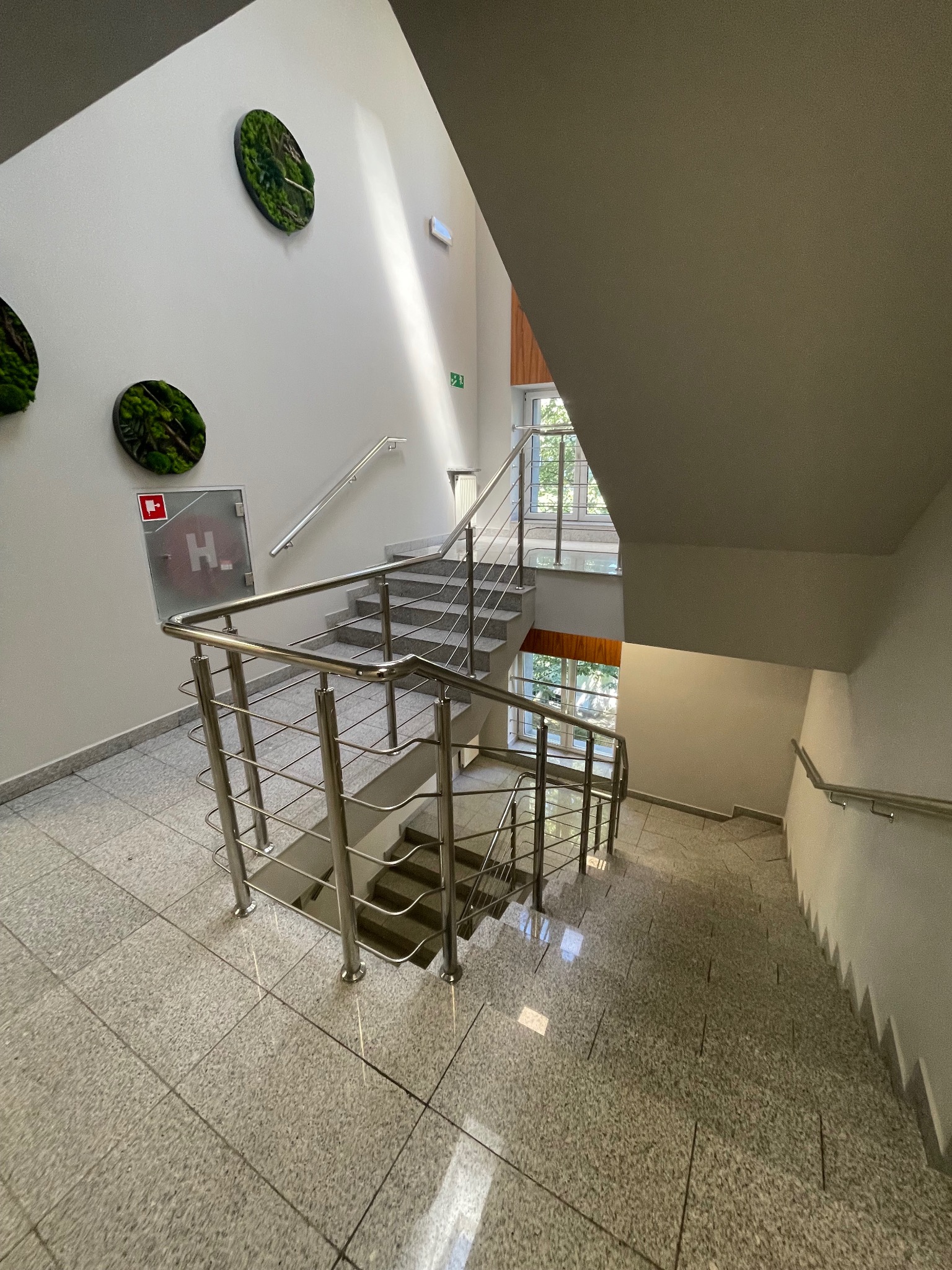 Jasne wnętrze klatki schodowej z metalowymi barierkami, okrągłymi dekoracjami roślinnymi na ścianie i szarą, wzorzystą podłogą z płytek.