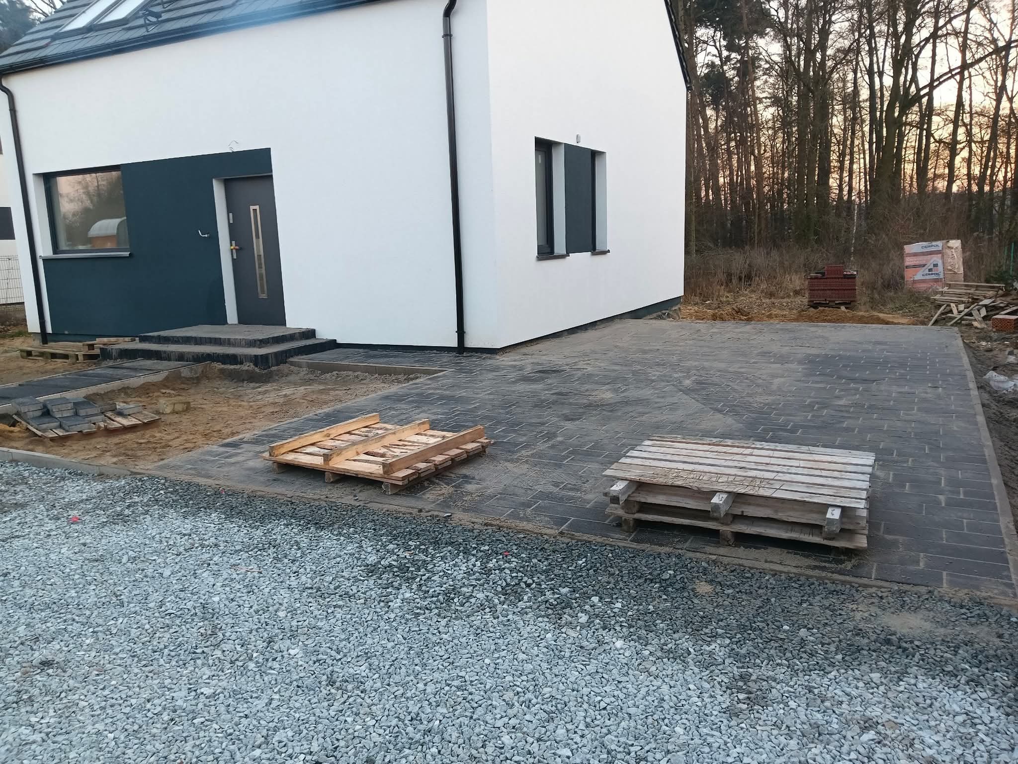 Nowoczesny dom z wybrukowanym podjazdem z szarej kostki. Częściowo ułożona kostka, palety z materiałem budowlanym. W tle las i zachodzące słońce.