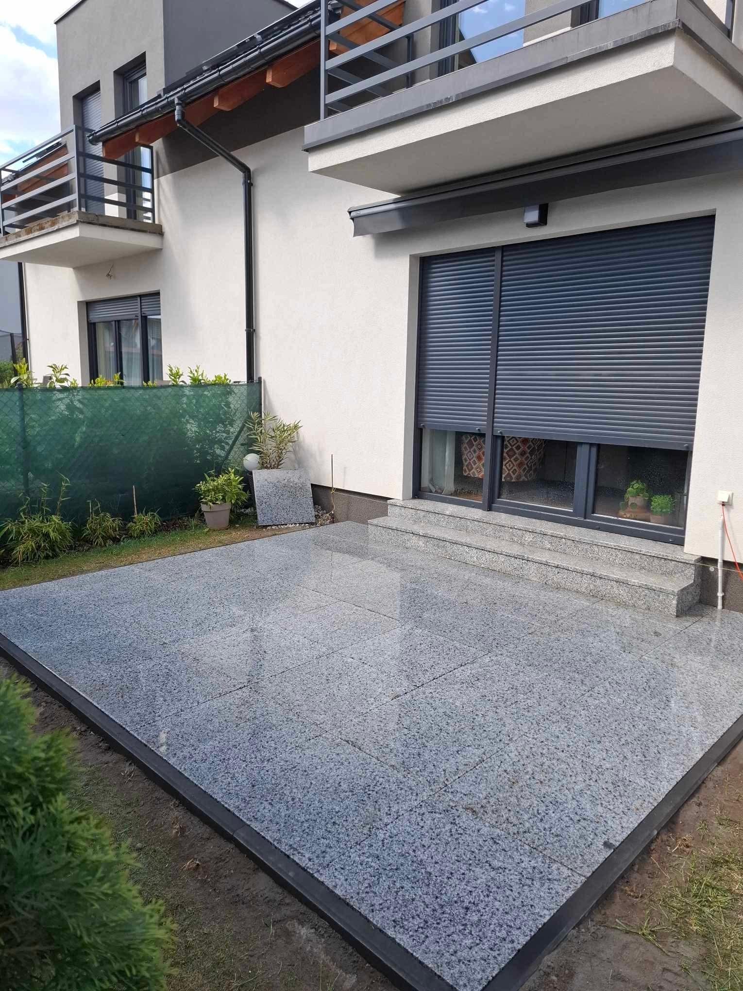 Granitowy taras przy domu z roletami zewnętrznymi. Widoczne rośliny w donicach i fragment zielonego ogrodzenia. Nowoczesne wykończenie.