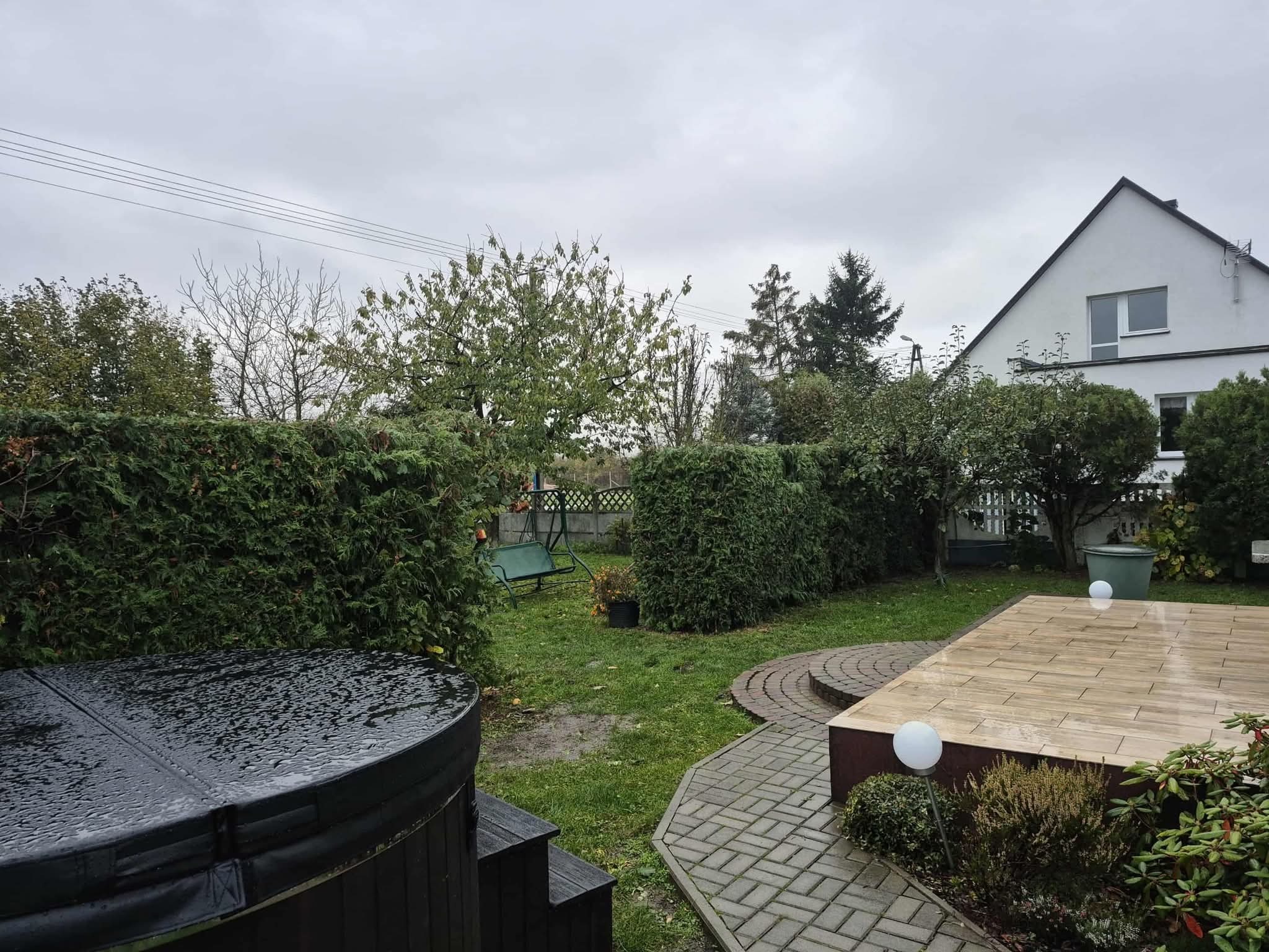 Jesienny ogród z jacuzzi, drewnianym tarasem i ścieżką z kostki. Widoczne tuje, trawnik i huśtawka. Na jacuzzi krople deszczu. Pochmurne niebo.