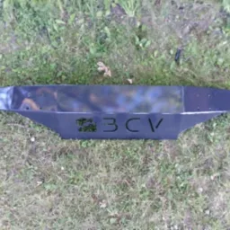 Czarny, metalowy element z wyciętym napisem '3CV' leżący na trawie.
