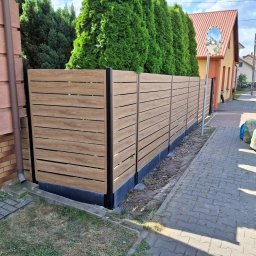 BCV SERWIS - MICHAŁ MAŁOLEPSZY - Nowoczesne ogrodzenie panelowe z poziomych desek w kolorze drewna, z czarnymi słupkami, oddziela posesję od chodnika. Za ogrodzeniem wysokie, zielone tuje.