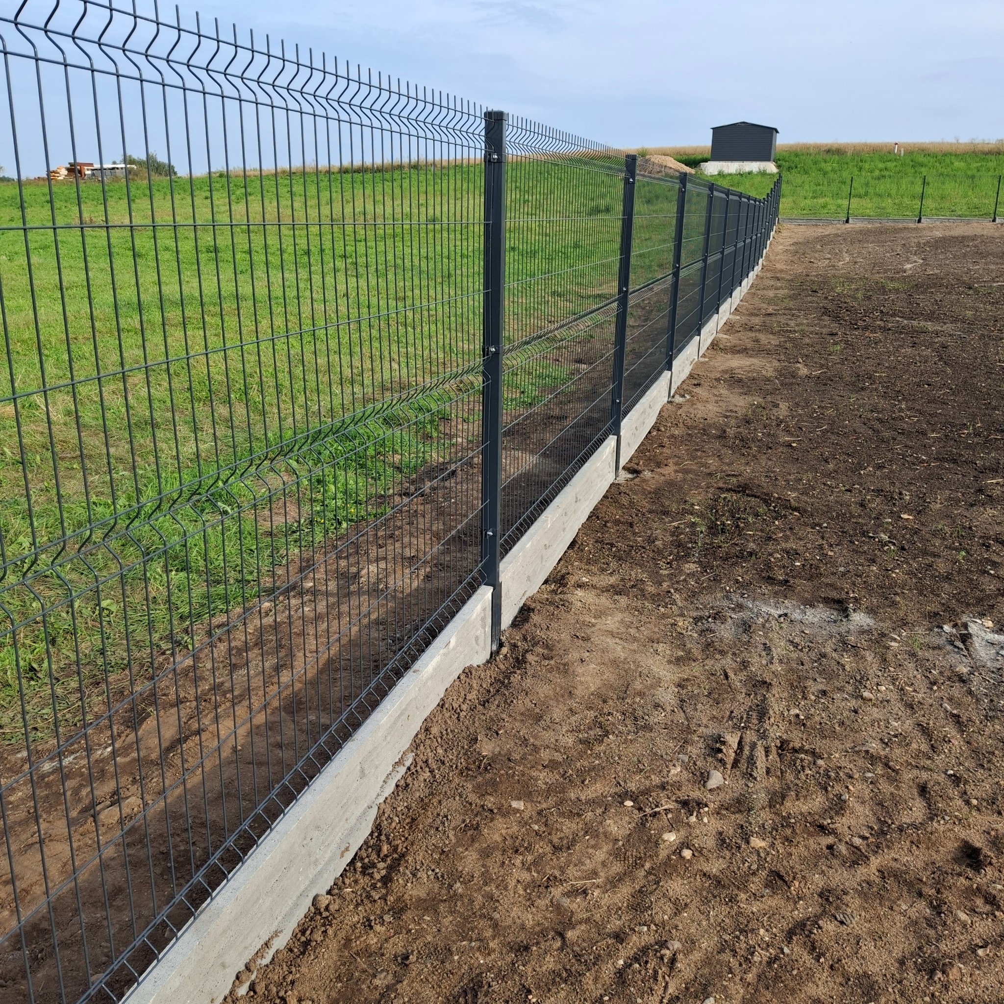 Nowoczesne ogrodzenie panelowe z podmurówką betonową, widok z boku na tle zieleni i nieba, prezentujące estetyczne i trwałe rozwiązanie ogradzające.