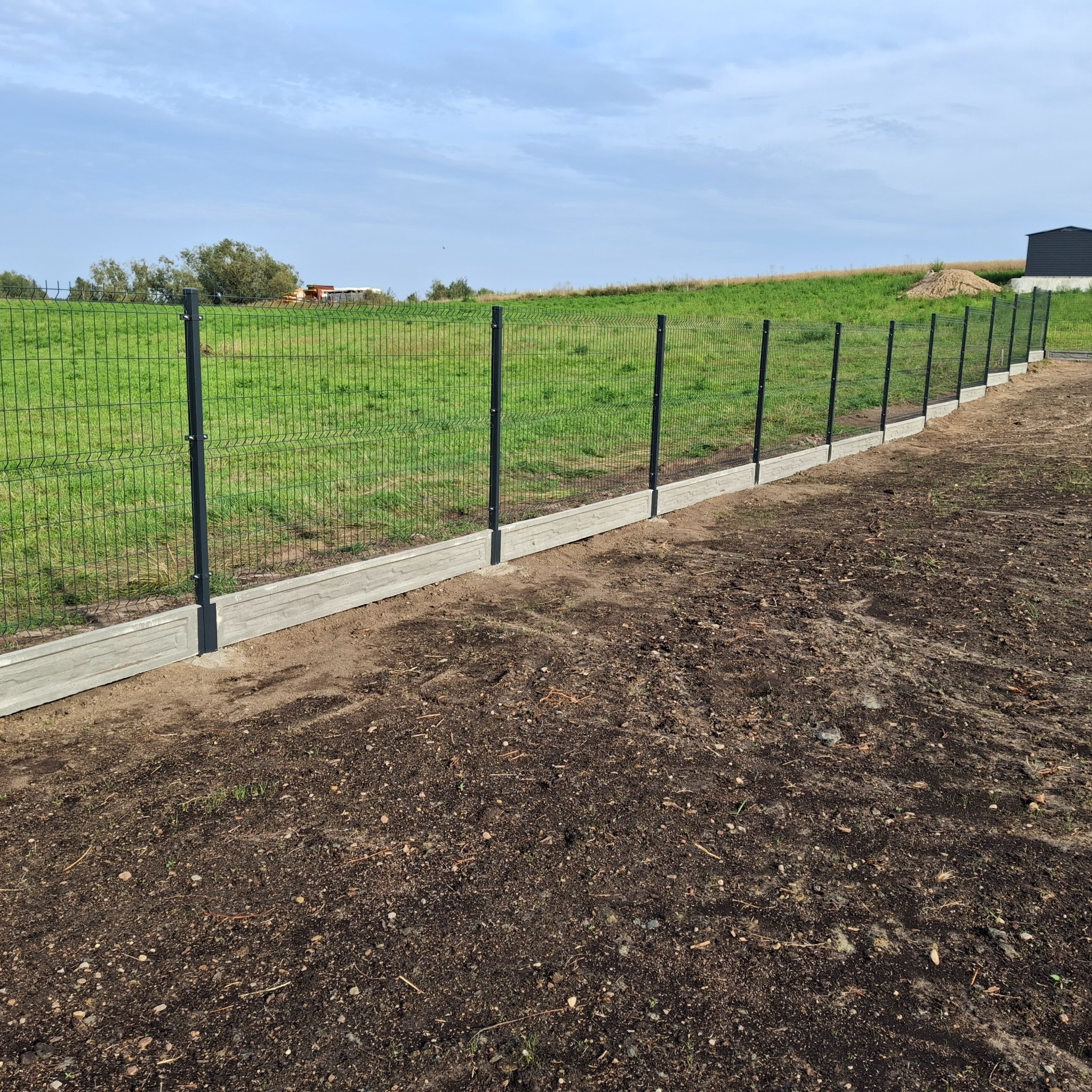 Nowoczesne ogrodzenie panelowe z podmurówką betonową na tle zielonego wzgórza i błękitnego nieba. Widoczny fragment posesji z budynkiem w oddali.