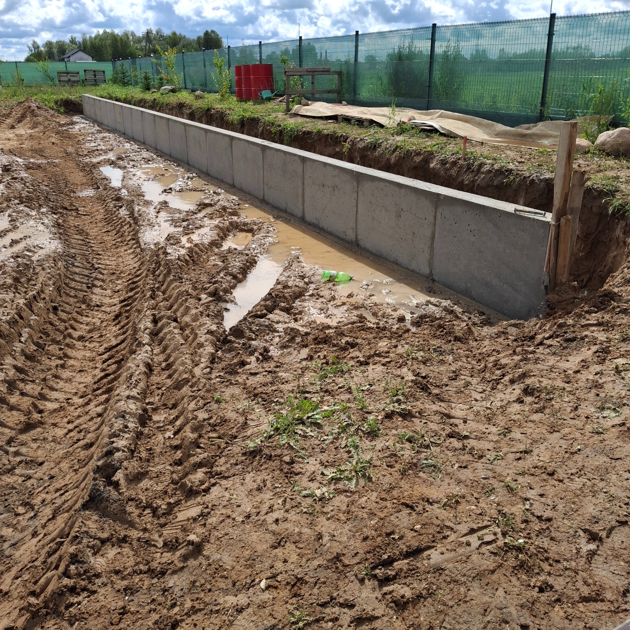 Betonowa podmurówka ogrodzenia w trakcie budowy, z widocznymi śladami opon w błotnistym terenie i fragmentem zielonego ogrodzenia siatkowego w tle.