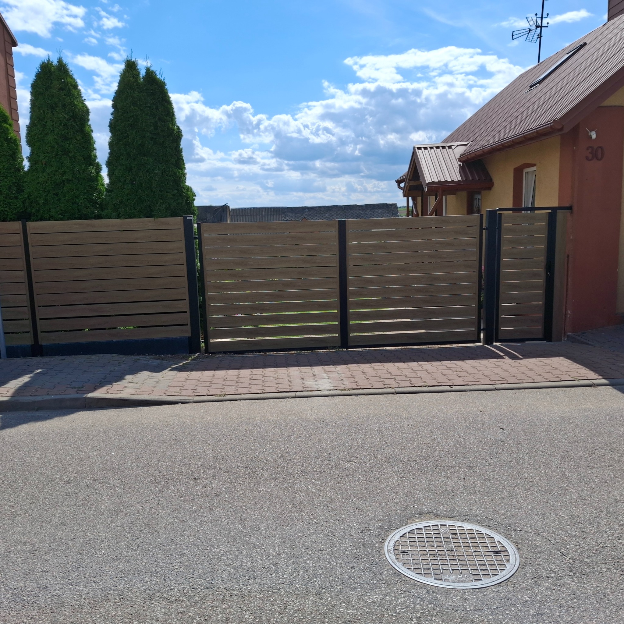 Nowoczesne, poziome ogrodzenie panelowe z metalowymi słupkami na tle domu z brązowym dachem i zielonymi tujami. Widok z poziomu ulicy, studzienka kanalizacyjna.