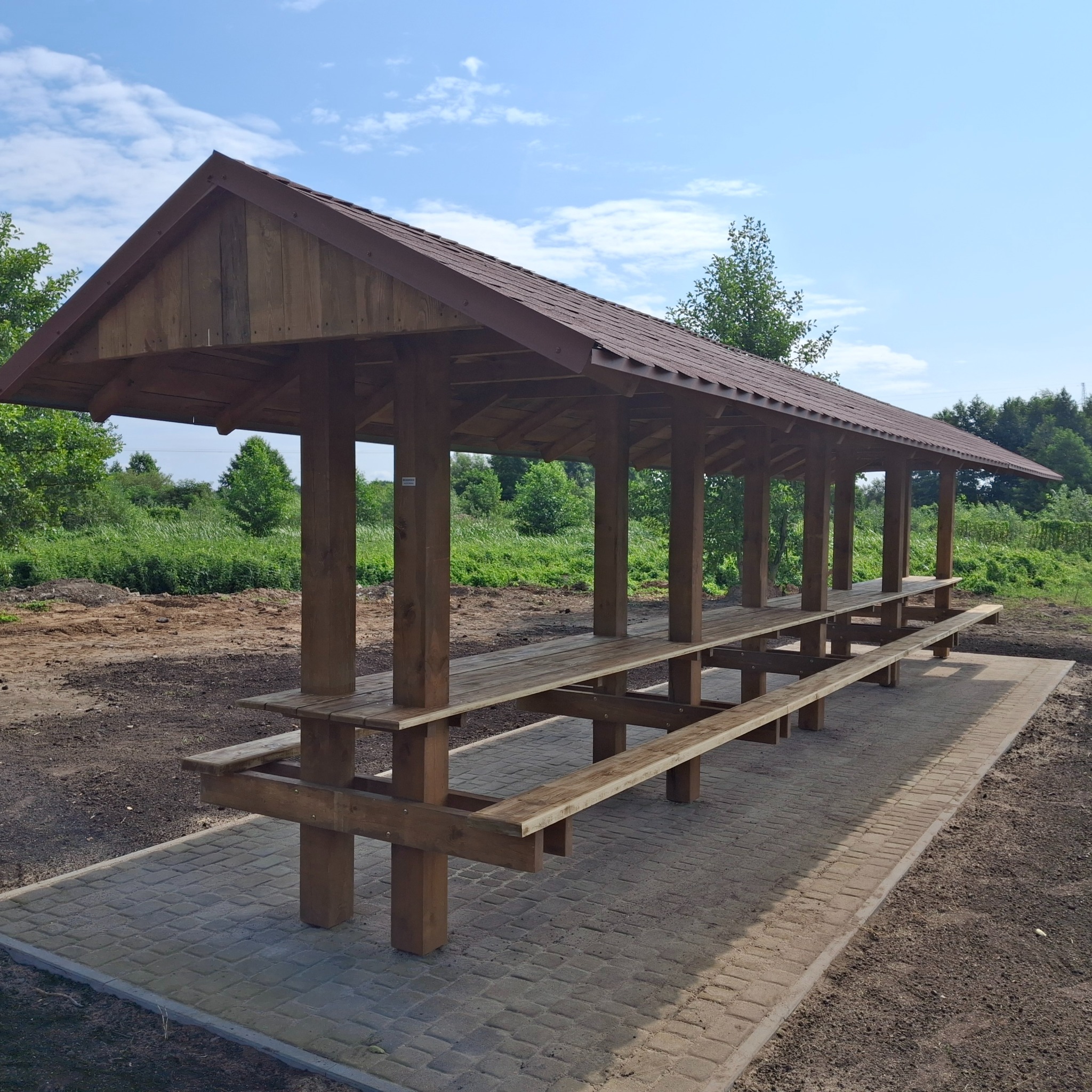 Drewniana pergola z długim stołem i ławkami na brukowanym podłożu, otoczona zielenią. Widok na konstrukcję w słoneczny dzień, z wyraźnymi detalami drewna i zadaszenia.