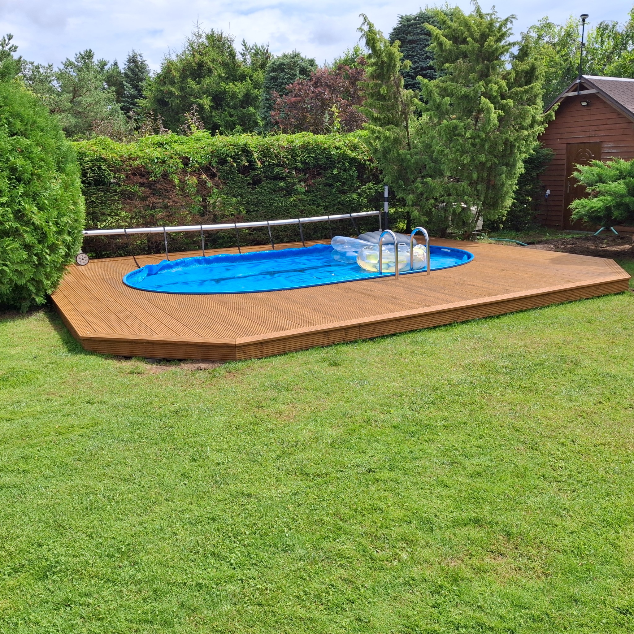 Drewniany taras otacza owalny, niebieski basen w ogrodzie. Widoczne drzewa iglaste i trawnik. Basen przykryty folią, obok drabinka i dmuchane zabawki.