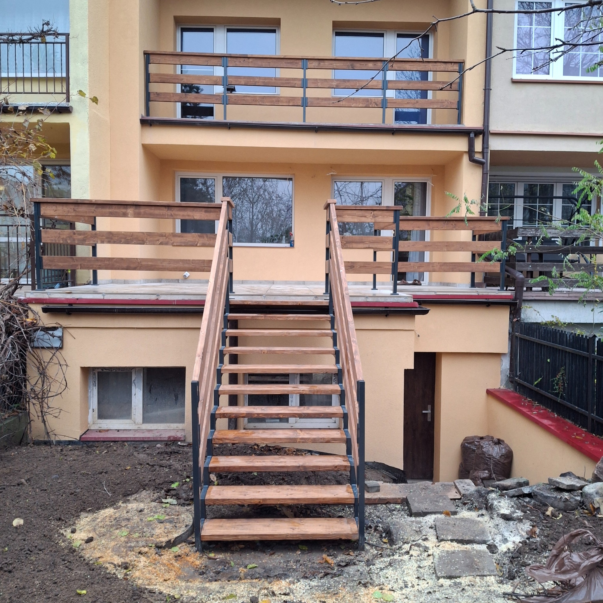 Nowoczesny taras z drewnianą balustradą i schodami z metalowymi elementami, prowadzącymi do ogrodu. Jasna elewacja budynku w tle. Widok z dołu.