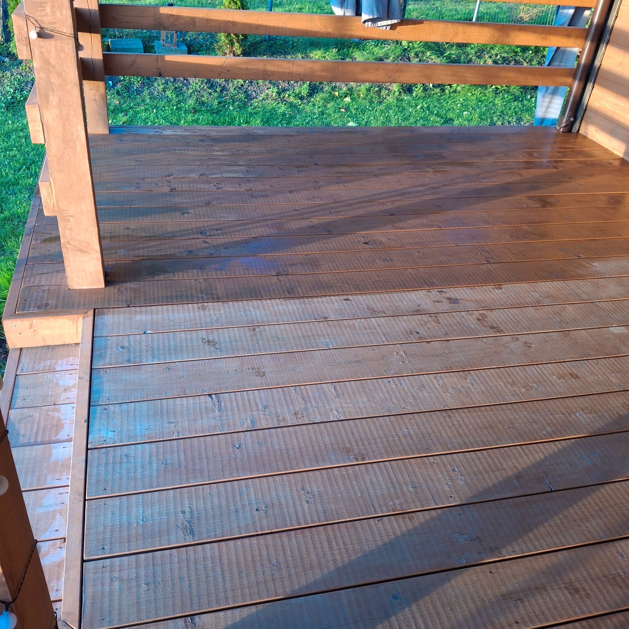 Impregnowany taras drewniany z balustradą, świeżo po malowaniu. Widoczne różnice w odcieniach drewna, podkreślające naturalną strukturę desek. Trawa w tle.