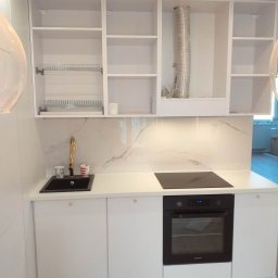 MARCIN MIROCHA - Jasna kuchnia z białymi szafkami, czarnym zlewem i złotą baterią, marmurowym backsplashiem i płytą indukcyjną, widoczne akcesoria kuchenne.