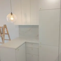 Nowoczesna kuchnia z białymi, matowymi frontami szafek, złotymi uchwytami i blatem z jasnego konglomeratu, oświetlona wiszącą lampą ze szklanym kloszem, z widoczną drabiną malarską w tle.