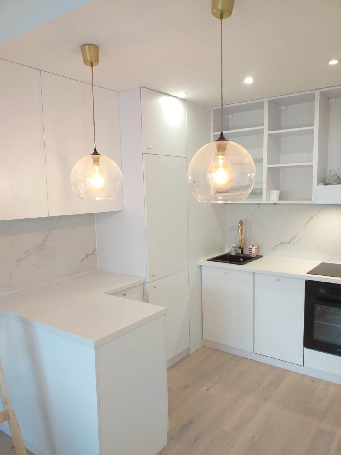 Nowoczesna, minimalistyczna kuchnia w bieli z drewnianą podłogą, wyposażona w okrągłe, szklane lampy wiszące z żarówkami Edisona, marmurowy backsplash i czarny zlew z złotą baterią.