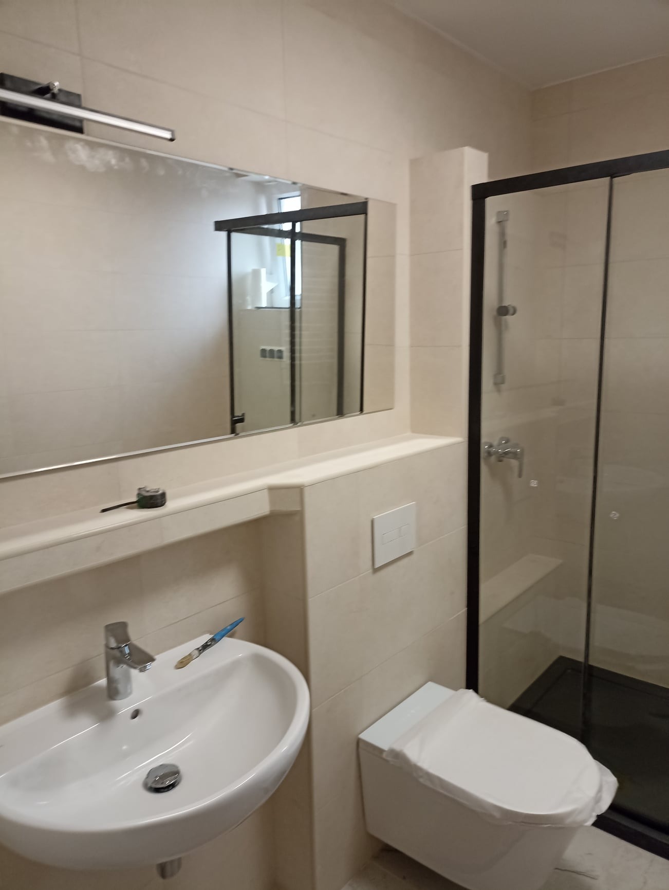 Jasna łazienka po remoncie: biała umywalka z baterią, toaleta z deską zabezpieczoną folią, kabina prysznicowa z czarną ramą, lustro z oświetleniem i miarka na półce.