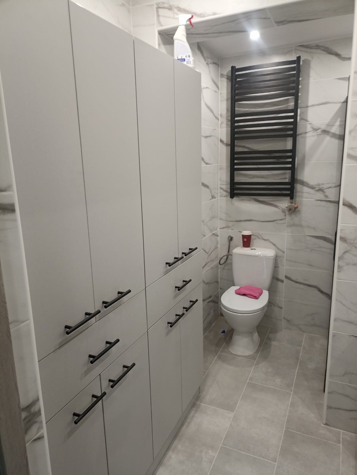 Jasnoszara łazienka z marmurowymi płytkami, szafkami z czarnymi uchwytami, grzejnikiem drabinkowym i toaletą z różowym ręcznikiem.