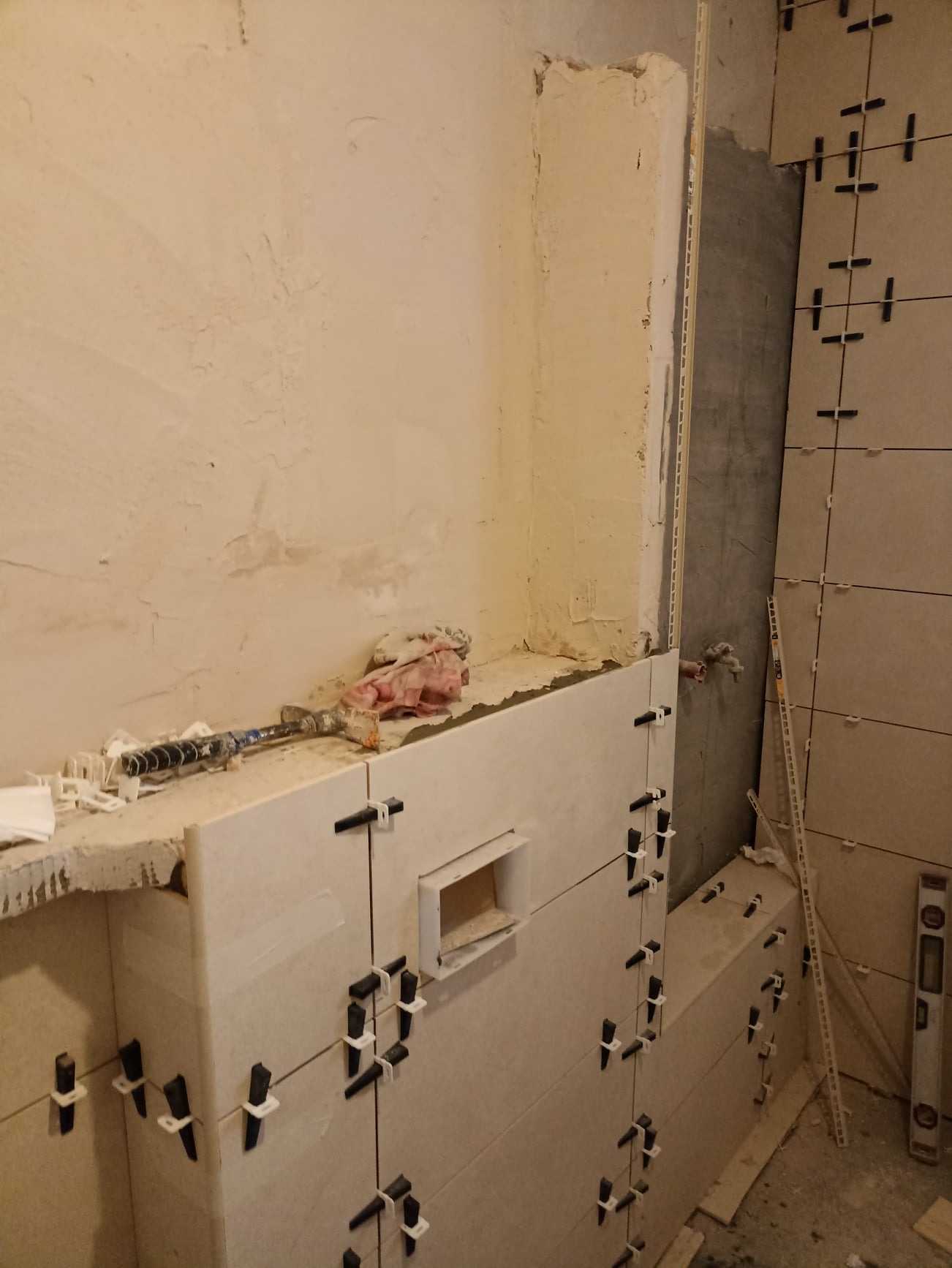 Montaż płytek ceramicznych w łazience, widoczne krzyżyki dystansowe, poziomica i narzędzia, ściana przygotowana do dalszych prac.