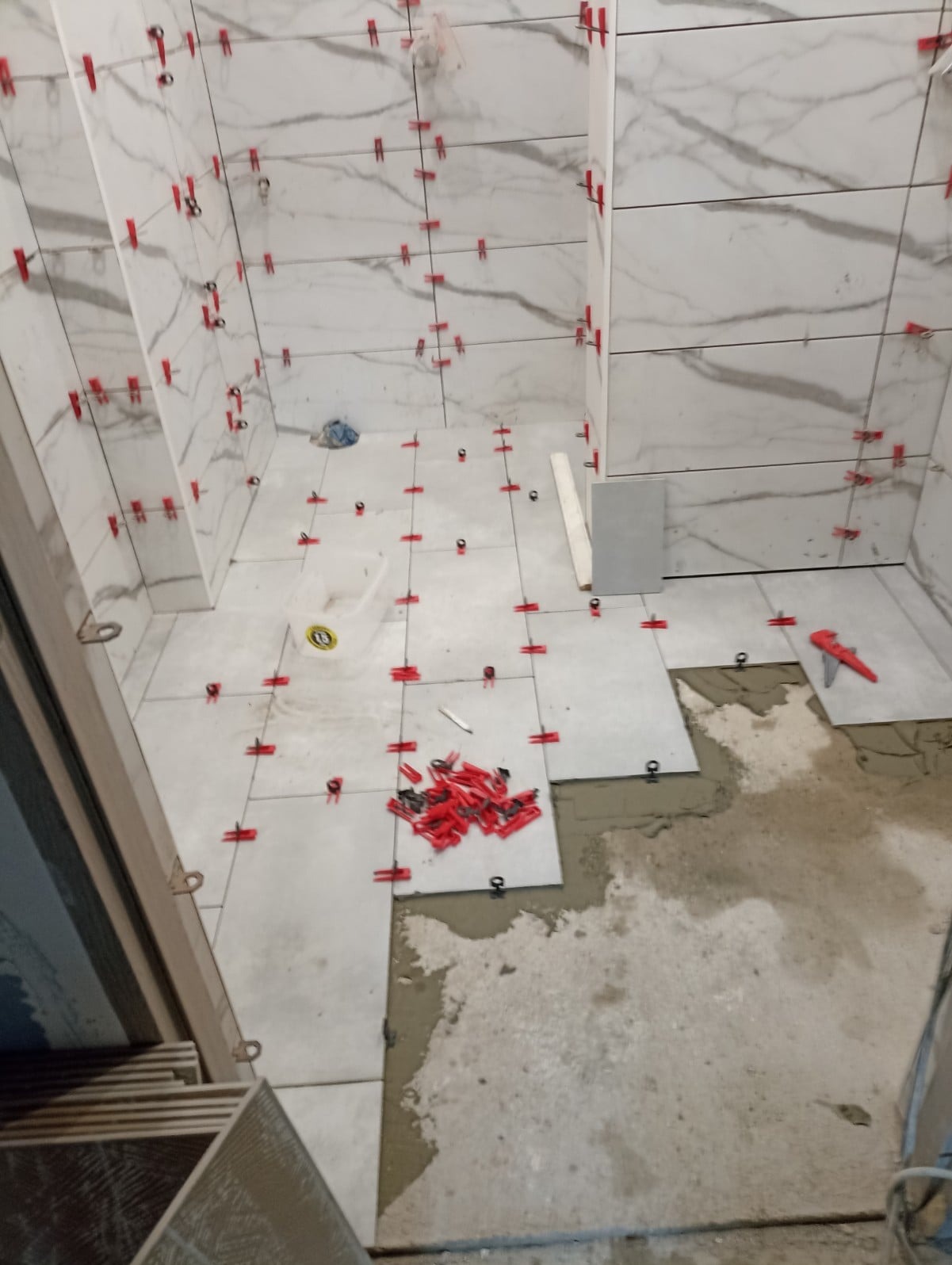 Układanie płytek imitujących marmur w łazience, widoczne czerwone klipsy poziomujące, zapas płytek i narzędzia.