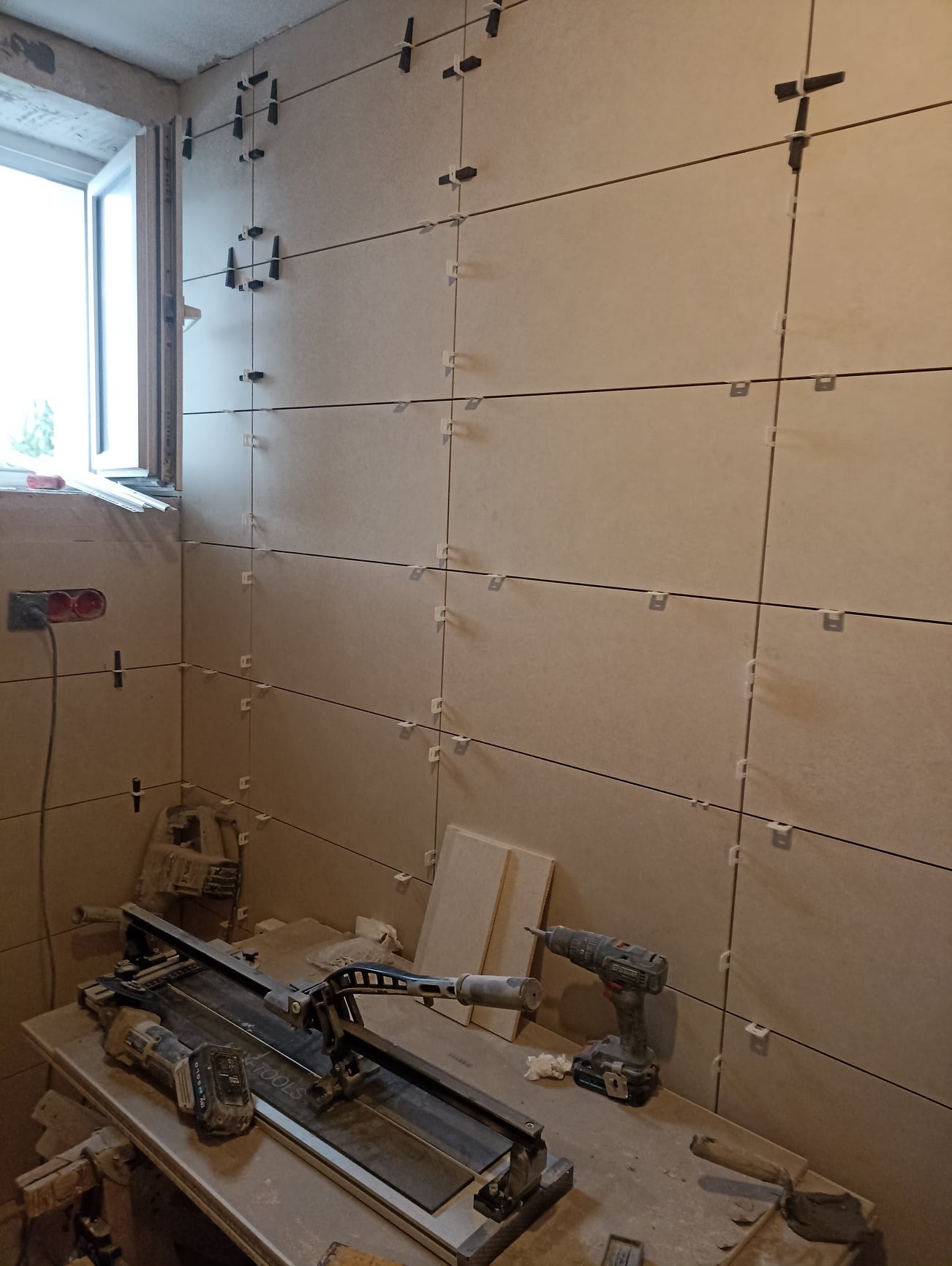 Ściana łazienki w trakcie układania płytek ceramicznych, widoczne krzyżyki dystansowe, narzędzia glazurnicze: przecinarka do płytek i wiertarko-wkrętarka.