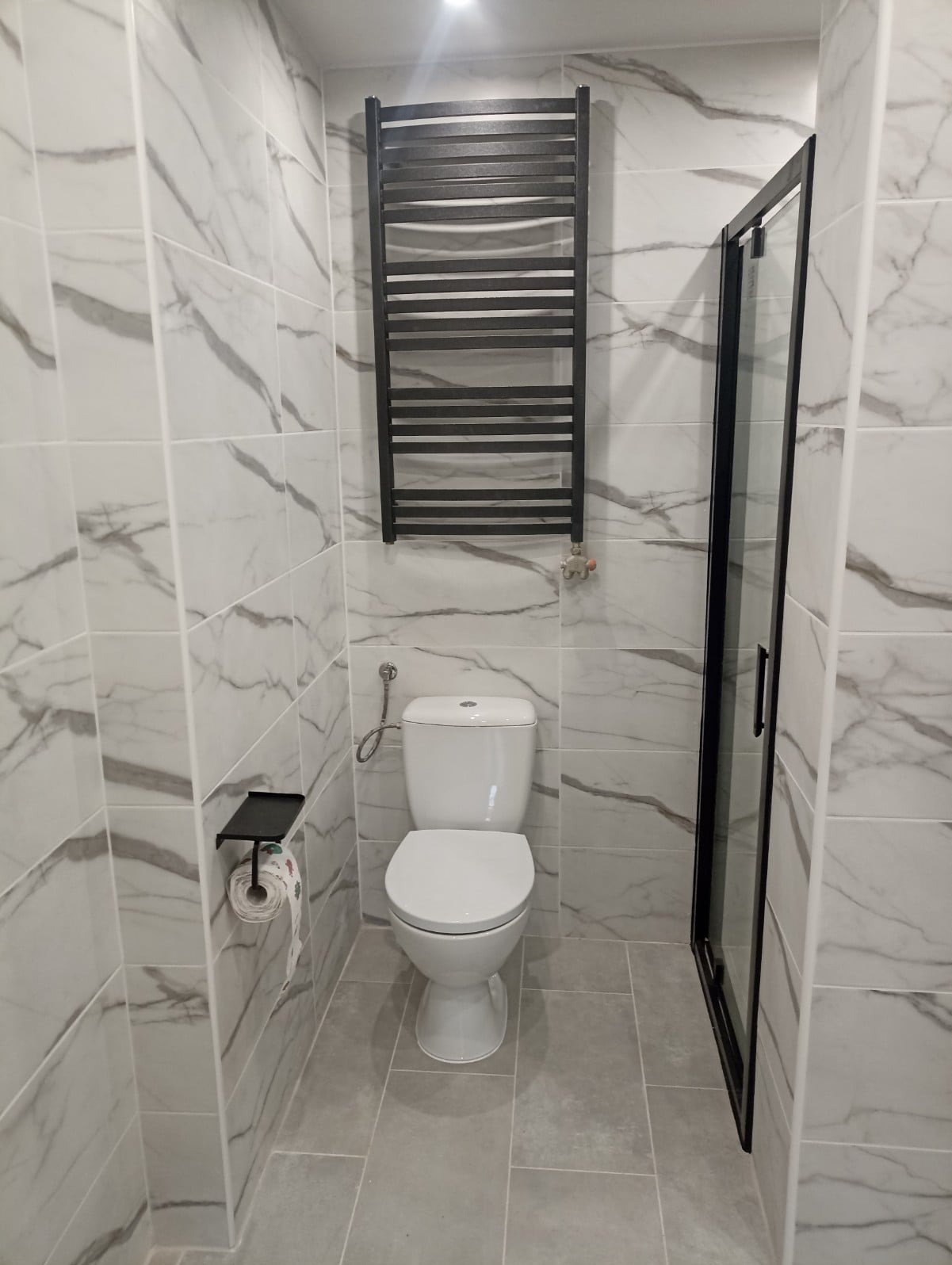 Nowoczesna łazienka z marmurowymi płytkami, wiszącym grzejnikiem drabinkowym w kolorze czarnym, białą toaletą i kabiną prysznicową z czarną ramą.